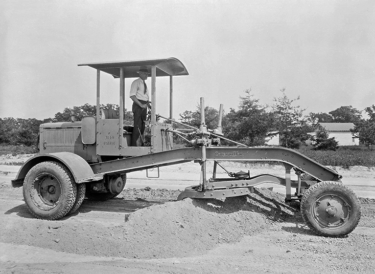 Ιστορική φωτογραφία του πρώτου Caterpillar Auto Patrol, μηχανικού ισοπεδωτή του 1931, με τον χειριστή πάνω στη μηχανή και λεπίδα στο κέντρο.
