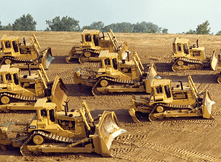 Σειρά από ερπυστριοφόρους προωθητές Caterpillar D11N με υπερυψωμένο οδοντωτό τροχό, παρατεταγμένοι σε εργοτάξιο με χωμάτινο υπόβαθρο.
